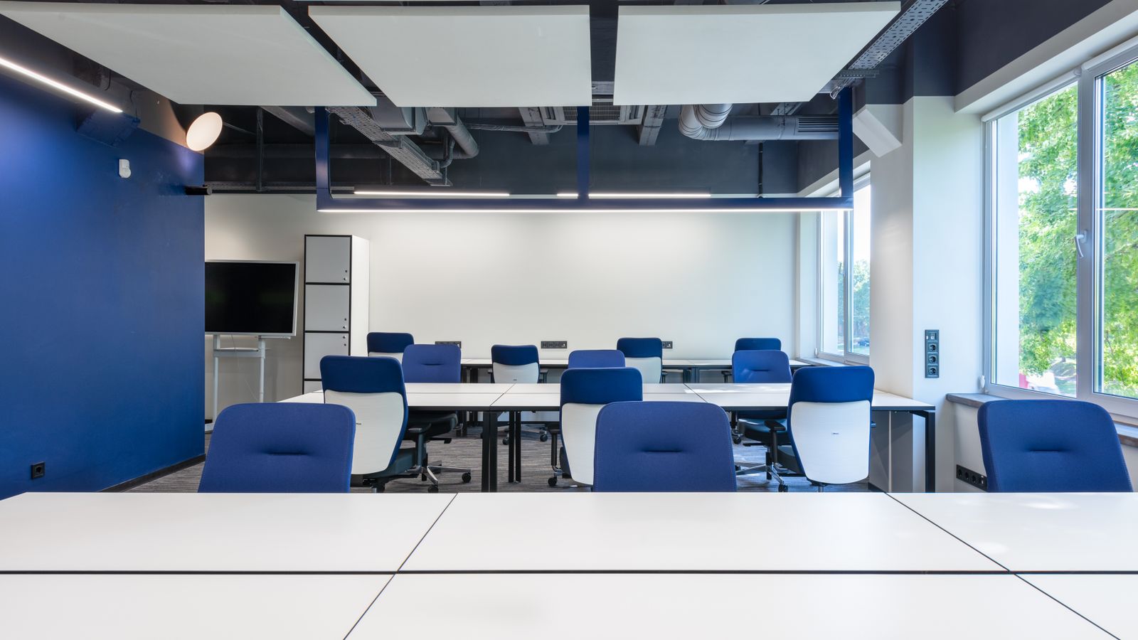 Various white desks with blue chairs placed in modern spacious workspace with big window and monitor in contemporary business center